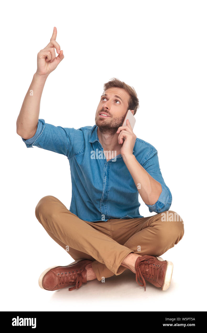 Seduto casual uomo parla al telefono e punti fino a qualcosa sopra la sua testa su sfondo bianco Foto Stock