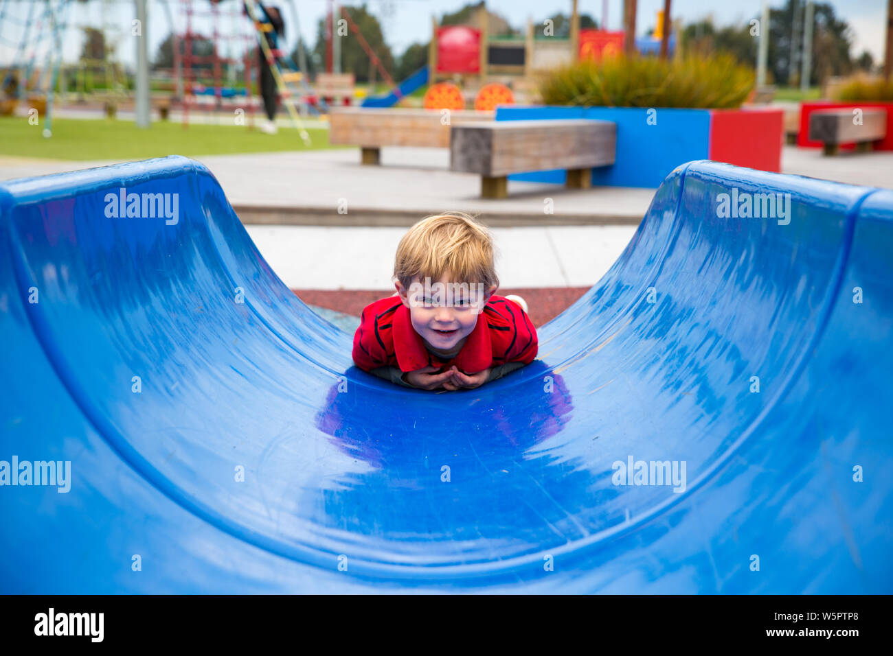 Due anni di vecchio ragazzo va in giù una slitta blu sulla sua pancia, divertirsi in un nuovo parco giochi, a Canterbury, Nuova Zelanda Foto Stock
