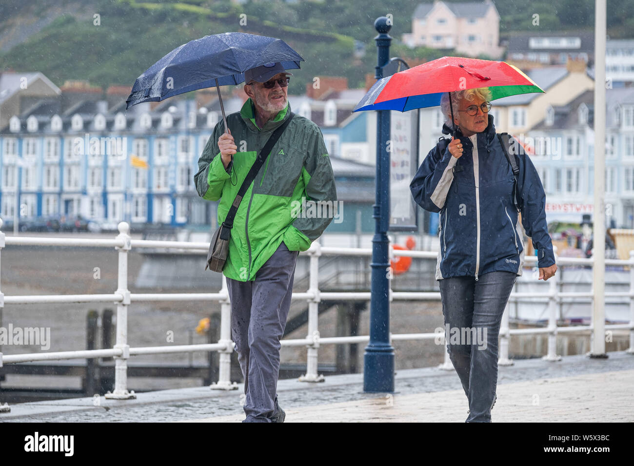 Aberystwyth, Wales, Regno Unito. Il 30 luglio 2019. I turisti nei loro impermeabili e ombrelli a piedi lungo la passeggiata in Aberystwyth sotto la pioggia come un nastro di thundery e umido molto tempo si diffonde attraverso molte delle parti centrali del Regno Unito, portando con sé il rischio di inondazione distruttiva e insidiose condizioni di guida. Credito Foto : Keith Morris/Alamy Live News Foto Stock