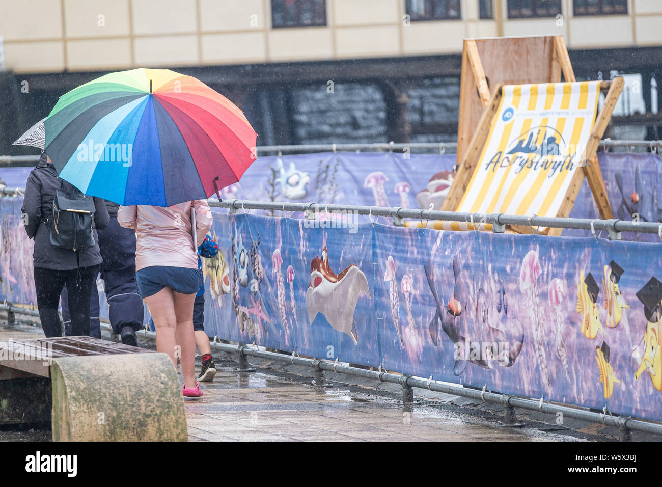 Aberystwyth, Wales, Regno Unito. Il 30 luglio 2019. I turisti nei loro impermeabili e ombrelli a piedi lungo la passeggiata in Aberystwyth sotto la pioggia come un nastro di thundery e umido molto tempo si diffonde attraverso molte delle parti centrali del Regno Unito, portando con sé il rischio di inondazione distruttiva e insidiose condizioni di guida. Credito Foto : Keith Morris/Alamy Live News Foto Stock