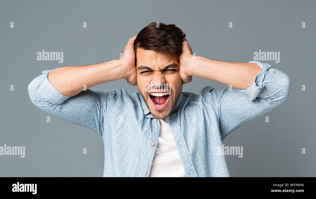 Emotivo uomo ispanico urlando che copre le sue orecchie su sfondo grigio Foto Stock