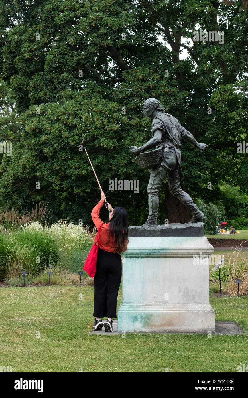 Femmina turistici asiatici di scattare una foto su un telefono intelligente del seminatore statua a kew gardens. Kew Gardens, Londra, Inghilterra Foto Stock