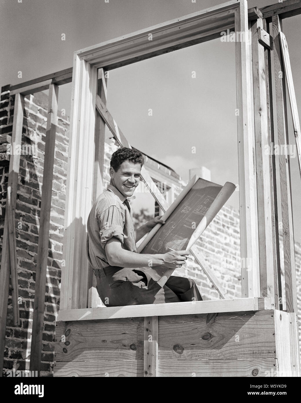 1940s sorridente giovane falegname OPERAIO EDILE seduto in casa incorniciate finestra lettura piani BLUEPRINT GUARDANDO TELECAMERA - b11688 HAR001 HARS GIOIA LIFESTYLE lavori falegname piani spazio copia di metà lunghezza fisica delle persone ispirazione maschi PROFESSIONE DI RISCHIO FIDUCIA B&W contatto visivo libertà successo gli obiettivi di occupazione di specialità felicità abilità Resistenza allegro ASSISTENZA CLIENTI carriera scelta eccitazione esterna orgoglio di progresso in occasione competente occupazioni sorrisi connessione con cornice concettuale gioiosa di precisione alla moda giovane uomo adulto BIANCO E NERO ETNIA CAUCASICA HAR001 Foto Stock
