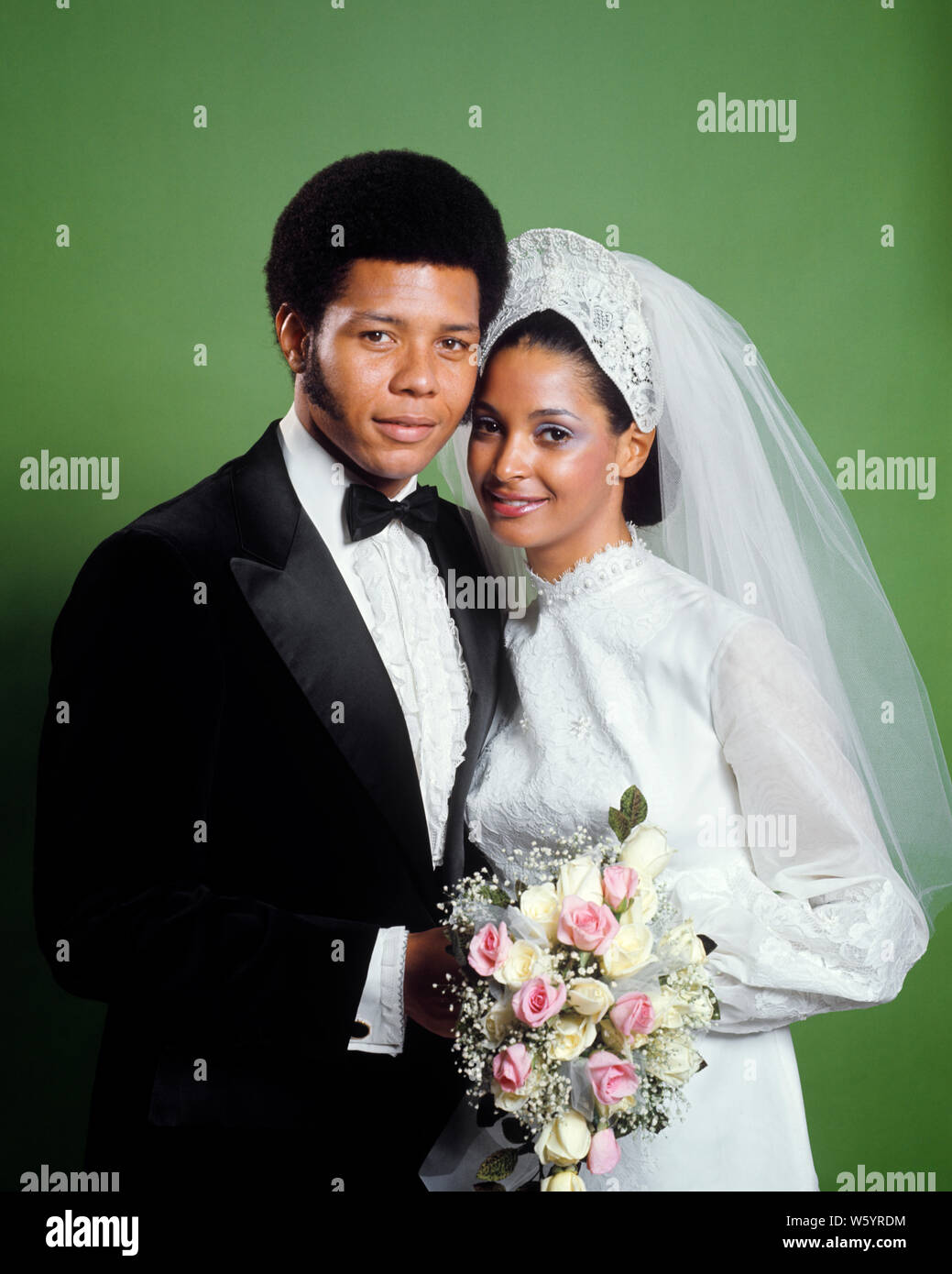 Anni Settanta ritratto sorridente AFRICAN-AMERICAN sposa e lo sposo uomo  che indossa lo smoking DONNA IN ABITO BIANCO VELO BIANCO ROSA bouquet di  rose - KB8763 HAR001 HARS giovane adulto lieta gioia