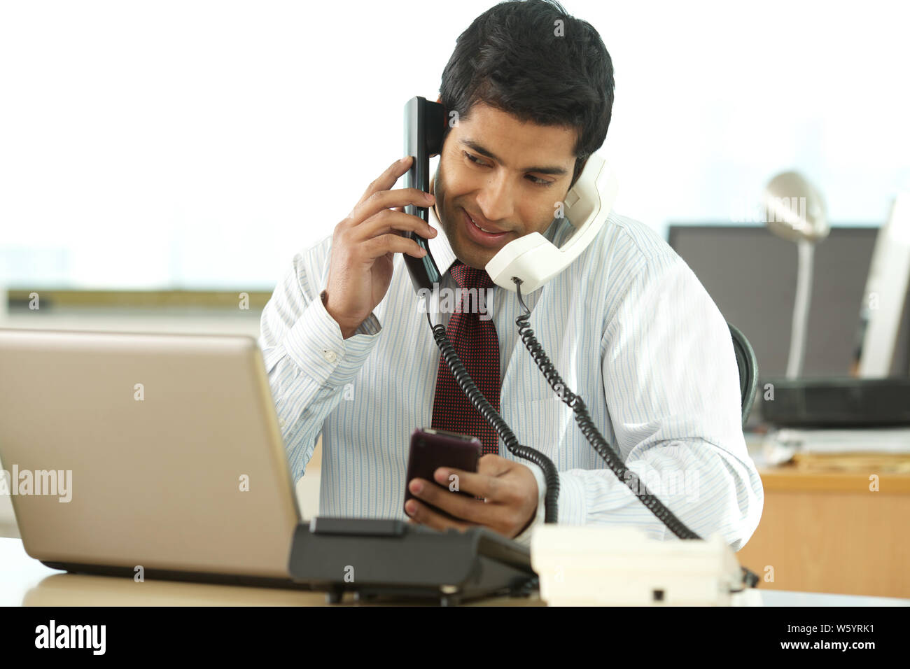 Uomo d'affari in ufficio con telefono cellulare e telefoni fissi a. allo stesso tempo Foto Stock