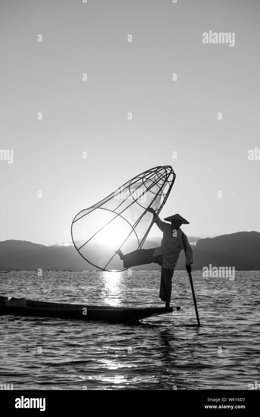 Immagine in bianco e nero del pescatore birmano silhouette in posa con una rete e un dispositivo di compressione al Lago Inle durante il tramonto, Myanmar Foto Stock