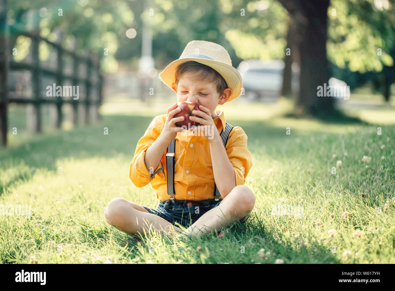 Carino adorabile ragazzo caucasica nel cappello di paglia di mangiare rosso  frutto di apple. Funny bambino in maglia gialla e Pantaloncini con bretelle  in parco con snack sani m Foto stock -