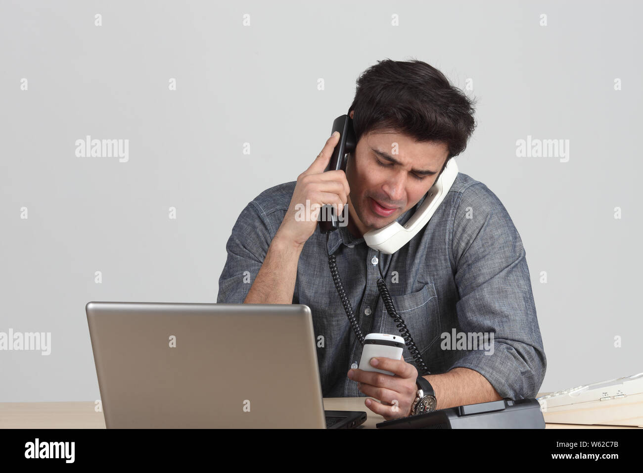 Uomo d'affari che mostra frustrazione durante l'uso di più telefoni e computer portatile Foto Stock