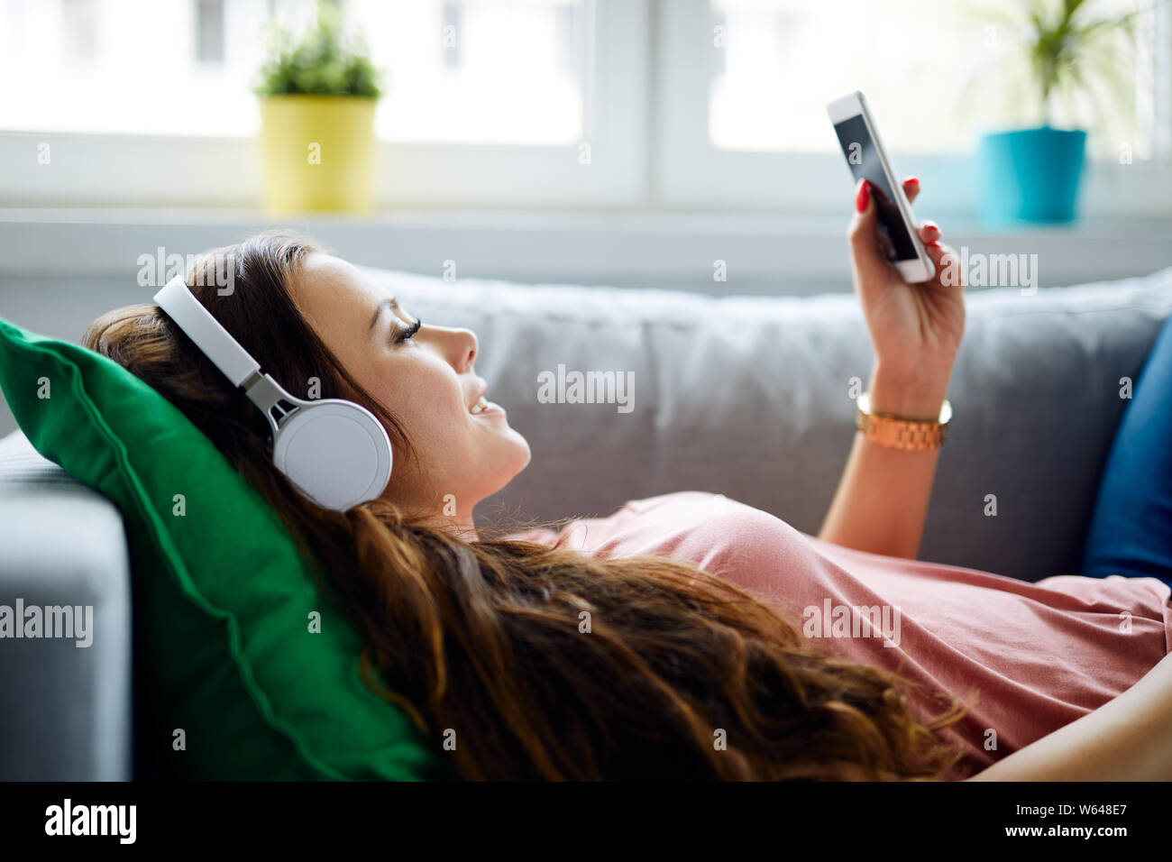 Vista laterale di una giovane donna rilassante sul divano di casa, l'ascolto di musica con cuffie e telefono azienda Foto Stock