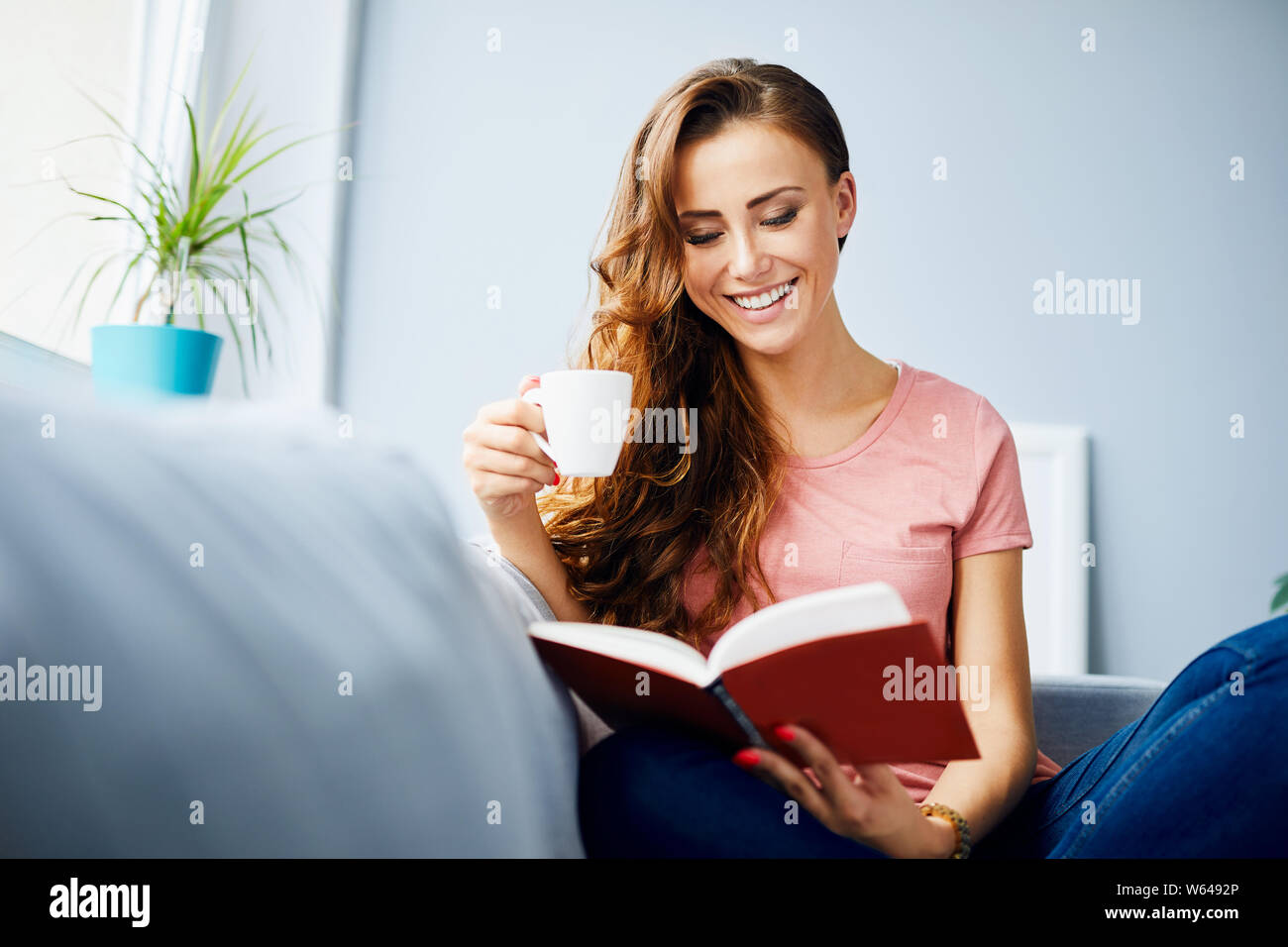 Piuttosto gioiosa giovane donna ridendo durante la lettura del libro e di bere caffè, rilassante sul divano di casa Foto Stock