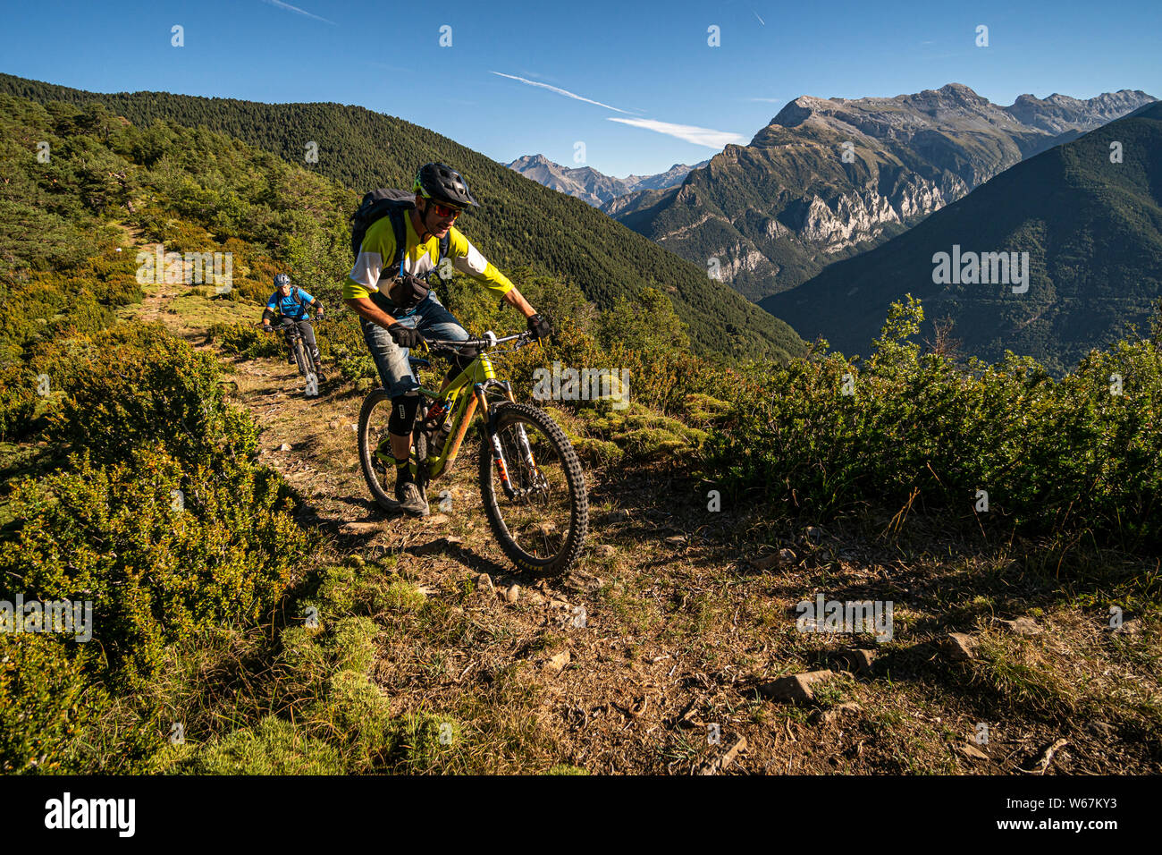 BIESCAS, HUESCA, Spagna. Due mountain bike a cavallo di un sentiero naturale attraverso la macchia aperta con picchi drammatici e cielo blu dietro. Foto Stock