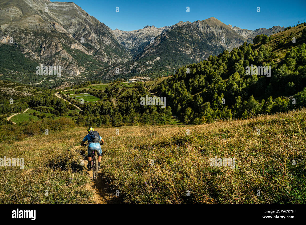BIESCAS, HUESCA, Spagna. Un mountain biker in sella a stretto sentiero attraverso prati aperti con picchi drammatici e cielo blu dietro. Foto Stock