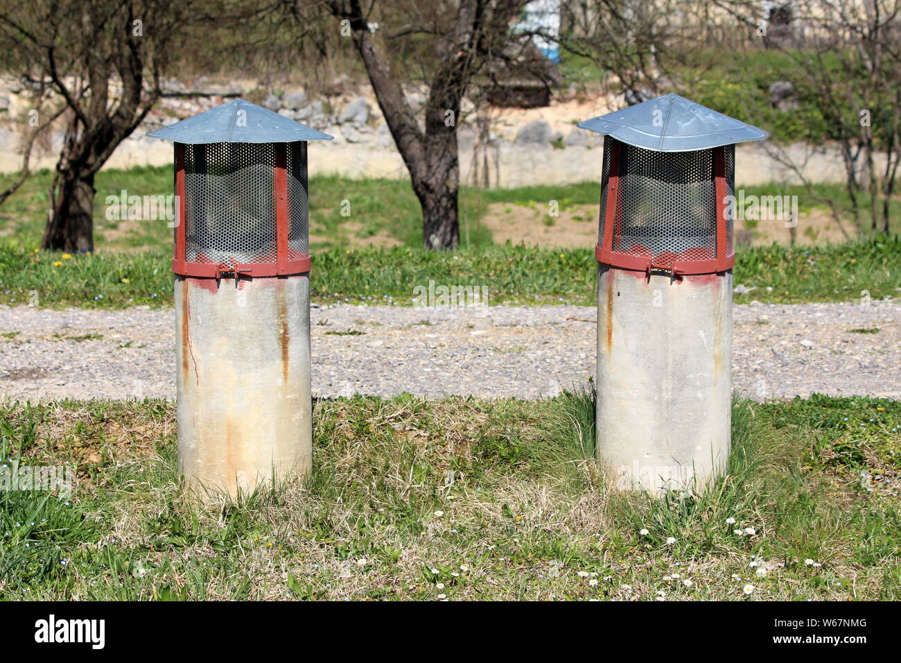 Due vecchio cemento tubi di ventilazione coperto con protezione di rete metallica circondato da erba non tagliata e ghiaia su caldo e soleggiato giorno di primavera Foto Stock