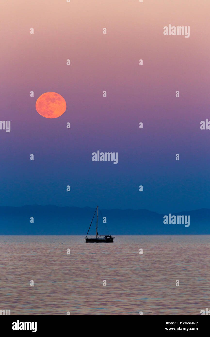 Rare Red Moon Rising oltre il mare mediterraneo in spagna con un pescatore della barca all'orizzonte Foto Stock