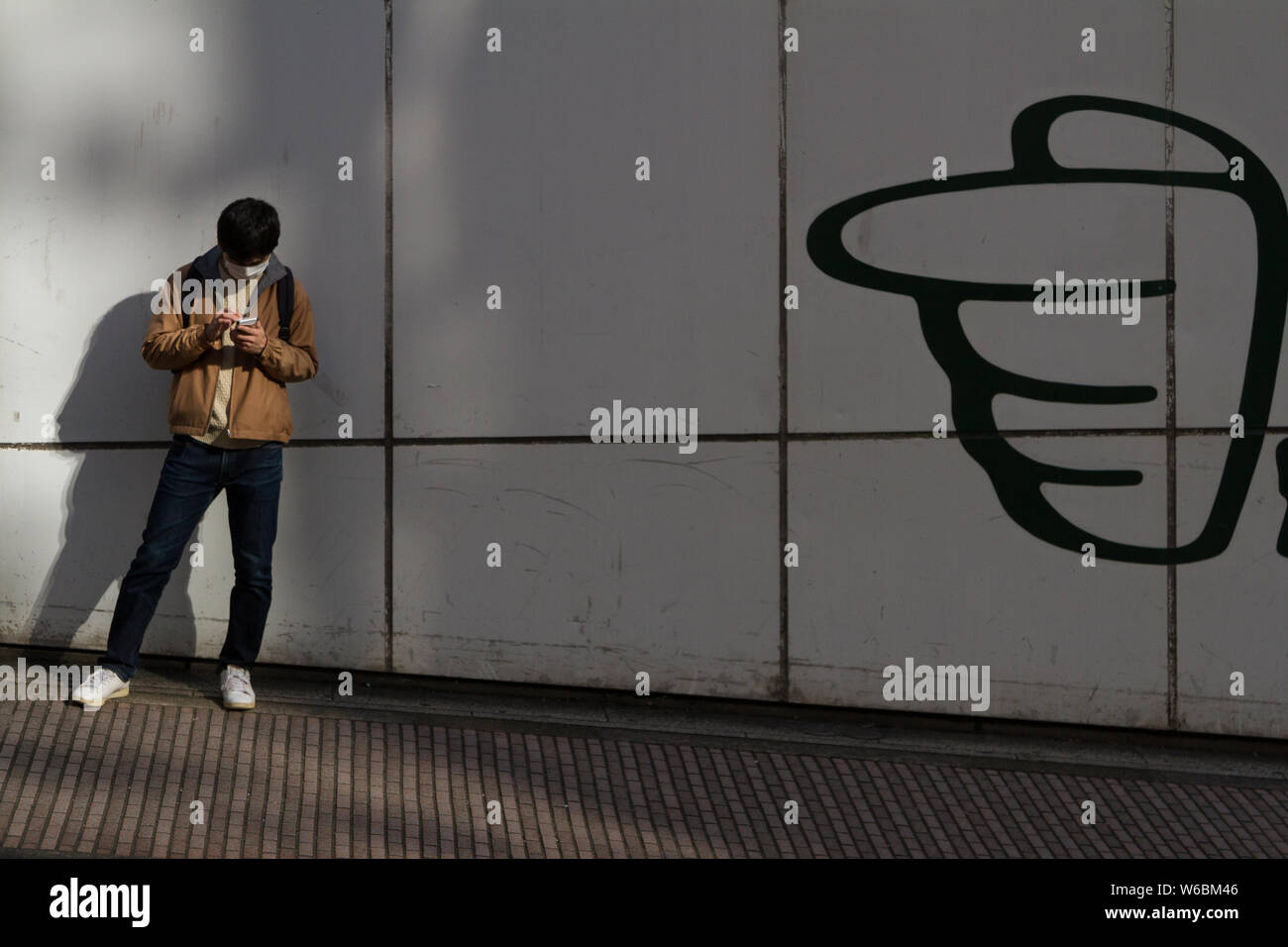 Un uomo giapponese controlla il suo smartphone accanto a una parete con un logo di una mano puntata su di esso. Shibuya, Tokyo, Giappone. Foto Stock