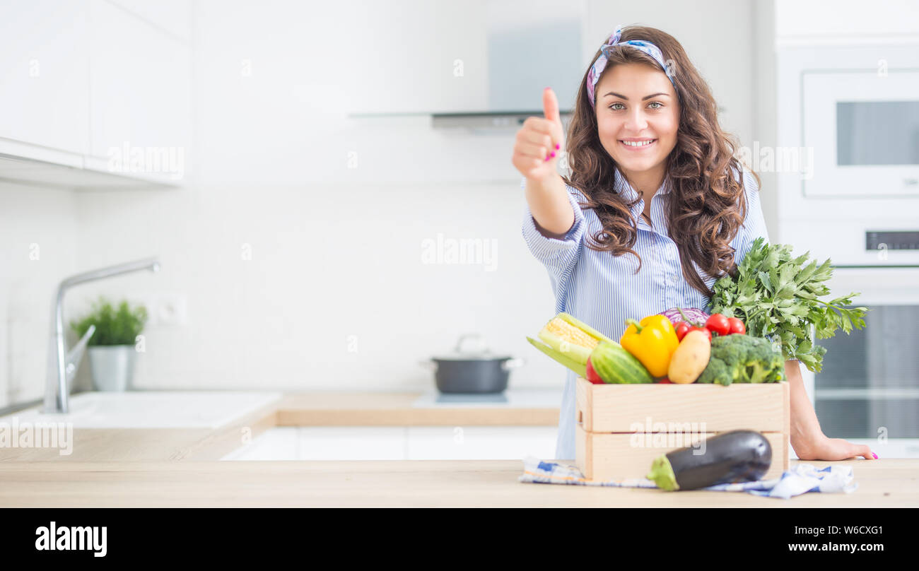 Donna nella sua cucina con scatola di legno piena di verdure biologiche che mostra Thumbs up. Foto Stock