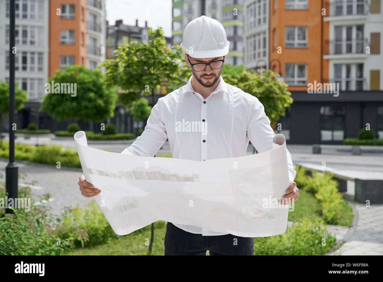 Giovane ingegnere in azienda hardhat costruzione architettonica di disegni su carta su sfondo del moderno sviluppo residenziale. Concetto di appartamenti nuovi edifici. Foto Stock