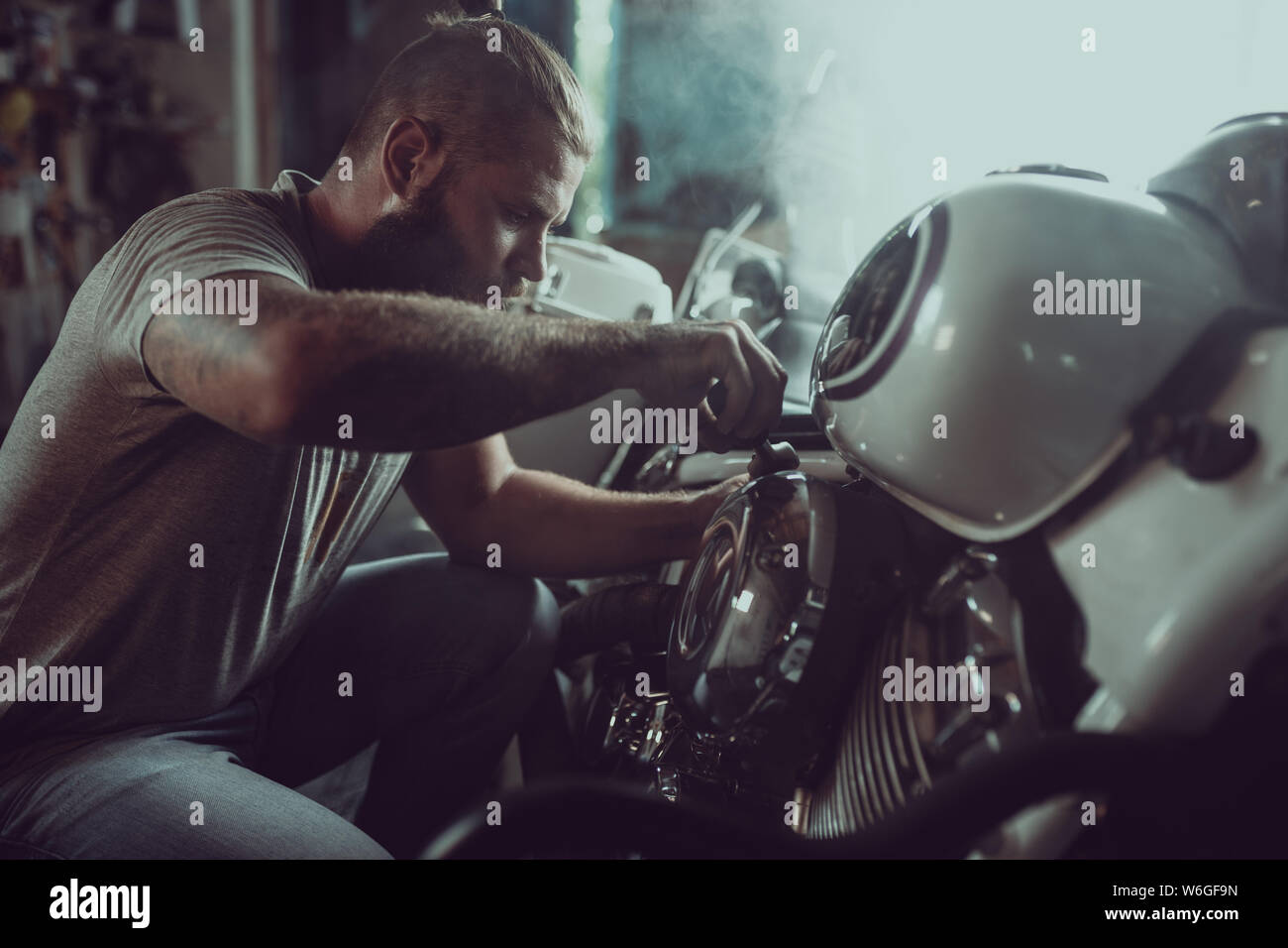 Bel uomo barbuto riparando il suo moto nel garage. Un uomo che indossa jeans e una maglietta. Foto Stock
