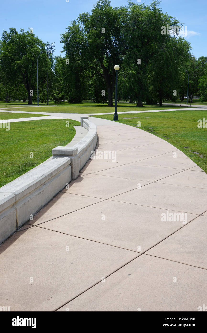 Un marciapiede in calcestruzzo si snoda attraverso un parco impostazione. Accanto al marciapiede è un basso muro di cemento. Ci sono grandi aree di erba e grandi alberi. Foto Stock