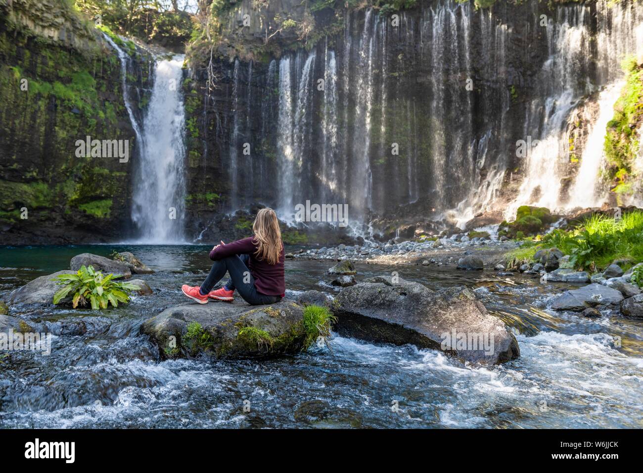 Giovane donna seduta su una pietra in un fiume, Shiraito cascata, Prefettura di Yamanashi, Giappone Foto Stock