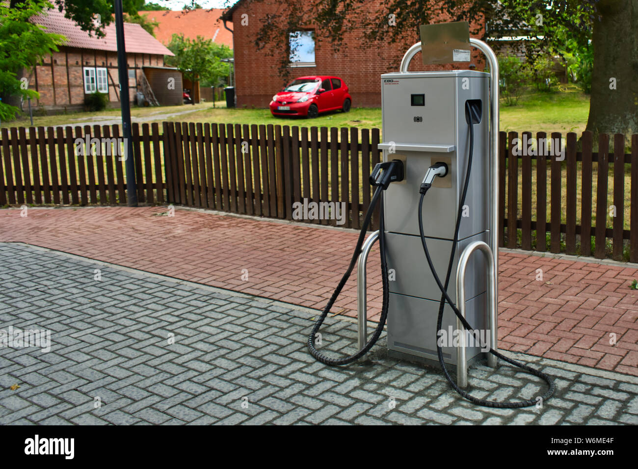 Gifhorn, Germania, Luglio 7, 2019: stazione di carica di un elettrico stazione di riempimento in un piccolo villaggio tedesco, con un recinto e una fattoria in background. Foto Stock