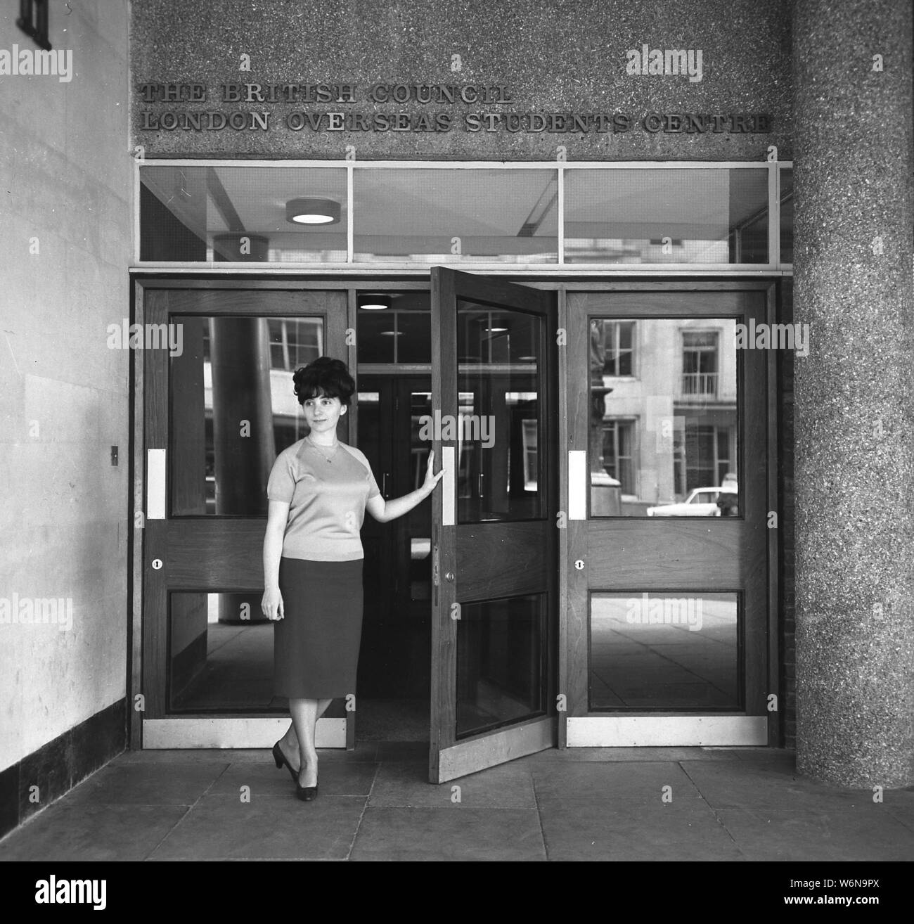 Anni sessanta, storico, una signora in piedi all'ingresso del London oltremare centro studenti del British Council, tenendo aperta la porta, forse in procinto di dare il benvenuto a nuovi arrivati gli studenti stranieri per l'edificio, Londra, Inghilterra, Regno Unito. Foto Stock