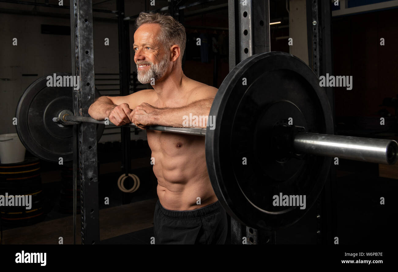 Ritratto di un affascinante vecchio atleta maschio con forti muscoli. L'atletico e sorridente uomo barbuto è appoggiata contro il barbell rack. Foto Stock