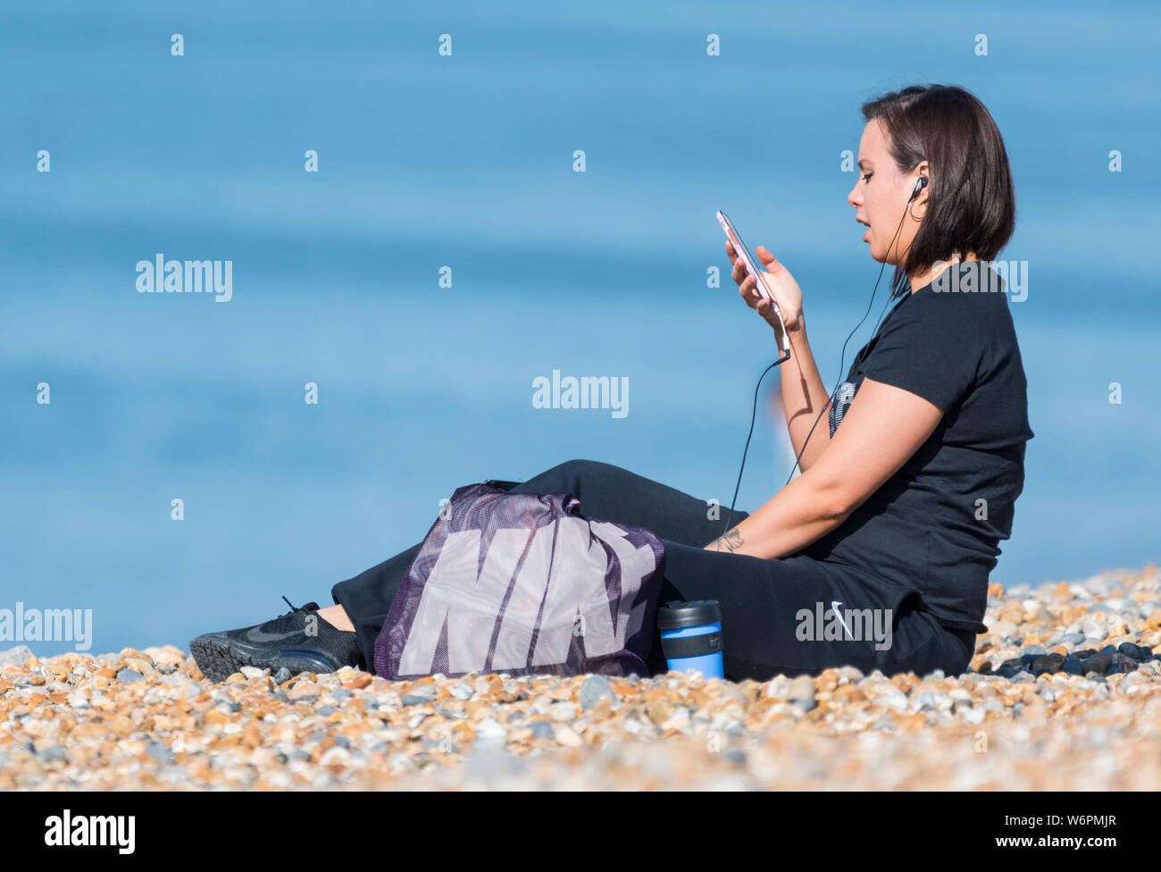 Vista laterale di una giovane donna seduta su una spiaggia con il suo smartphone con le cuffie per ascoltare musica. Utilizzando un telefono sulla spiaggia. Foto Stock