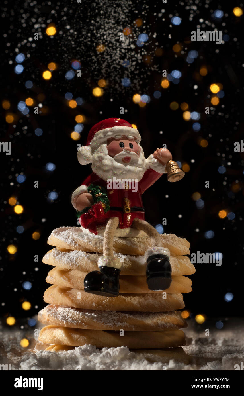 Babbo Natale seduto su una pila di ricco burro pezzata frollini con zucchero a velo con bokeh illuminazione effetto in background. Foto Stock