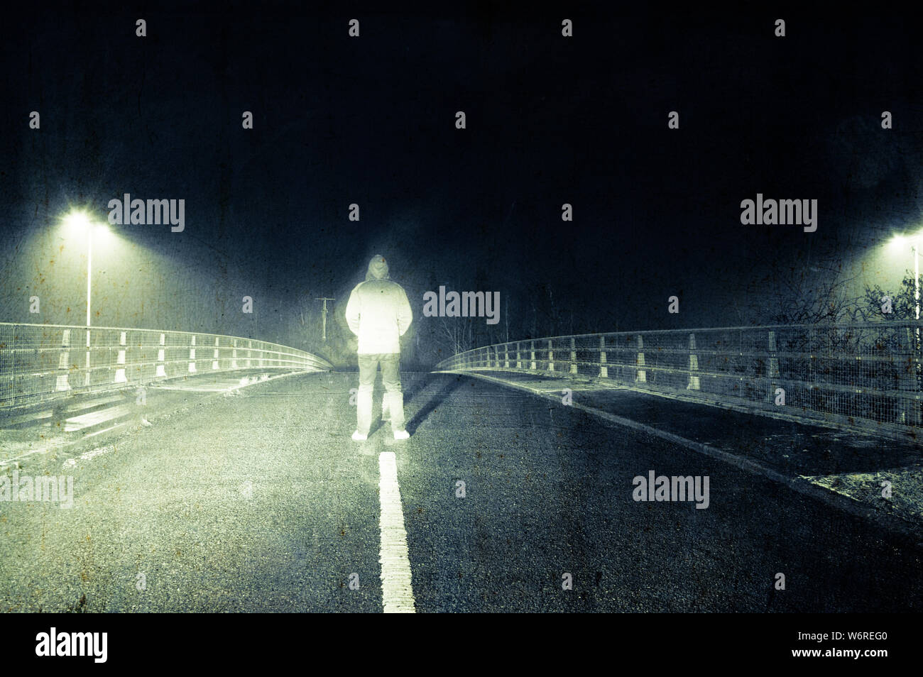 Un spooky incappucciati figura, in piedi nel mezzo di una strada di campagna su un inverni di notte. Con un vintage grunge modifica. Foto Stock