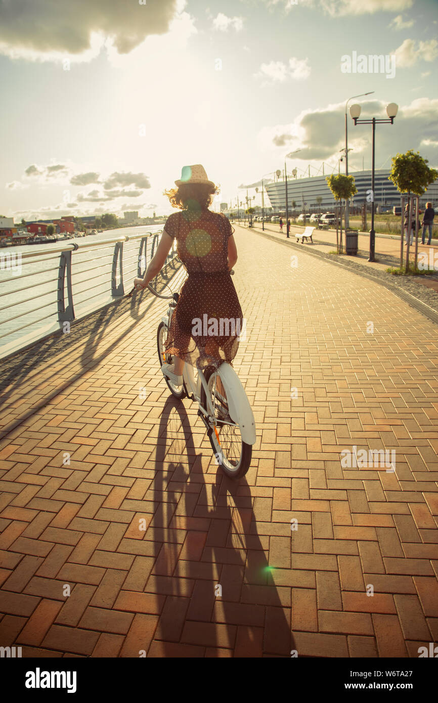 Giovani capelli ricci woman in hat e il vestito si muove in bicicletta  vicino al fiume in città, vista posteriore del tramonto Foto stock - Alamy