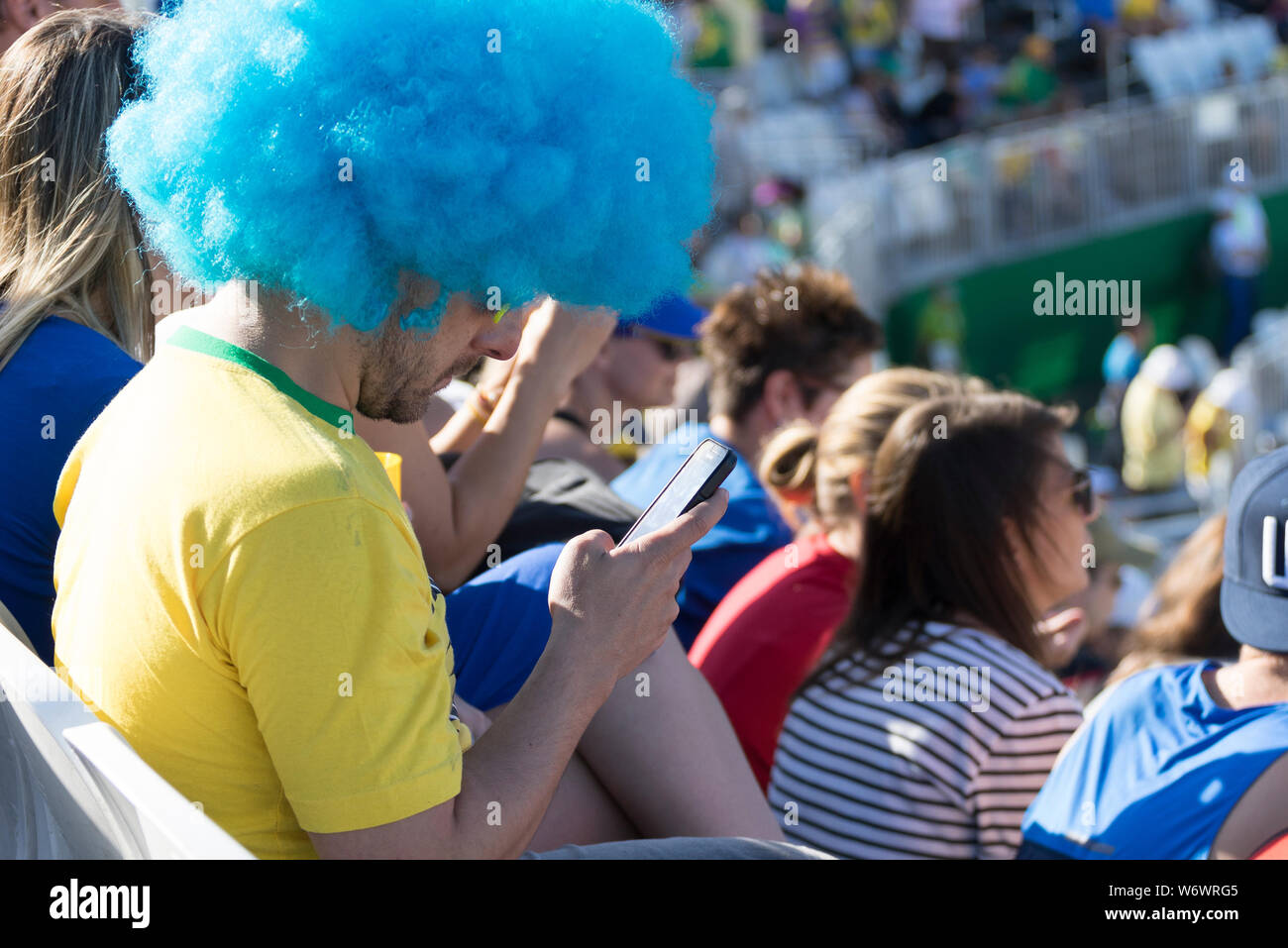 Sostenitore brasiliano con parrucca blu su guardando al suo telefono cellulare invece di ciò che sta succedendo allo stadio con la folla in background Foto Stock