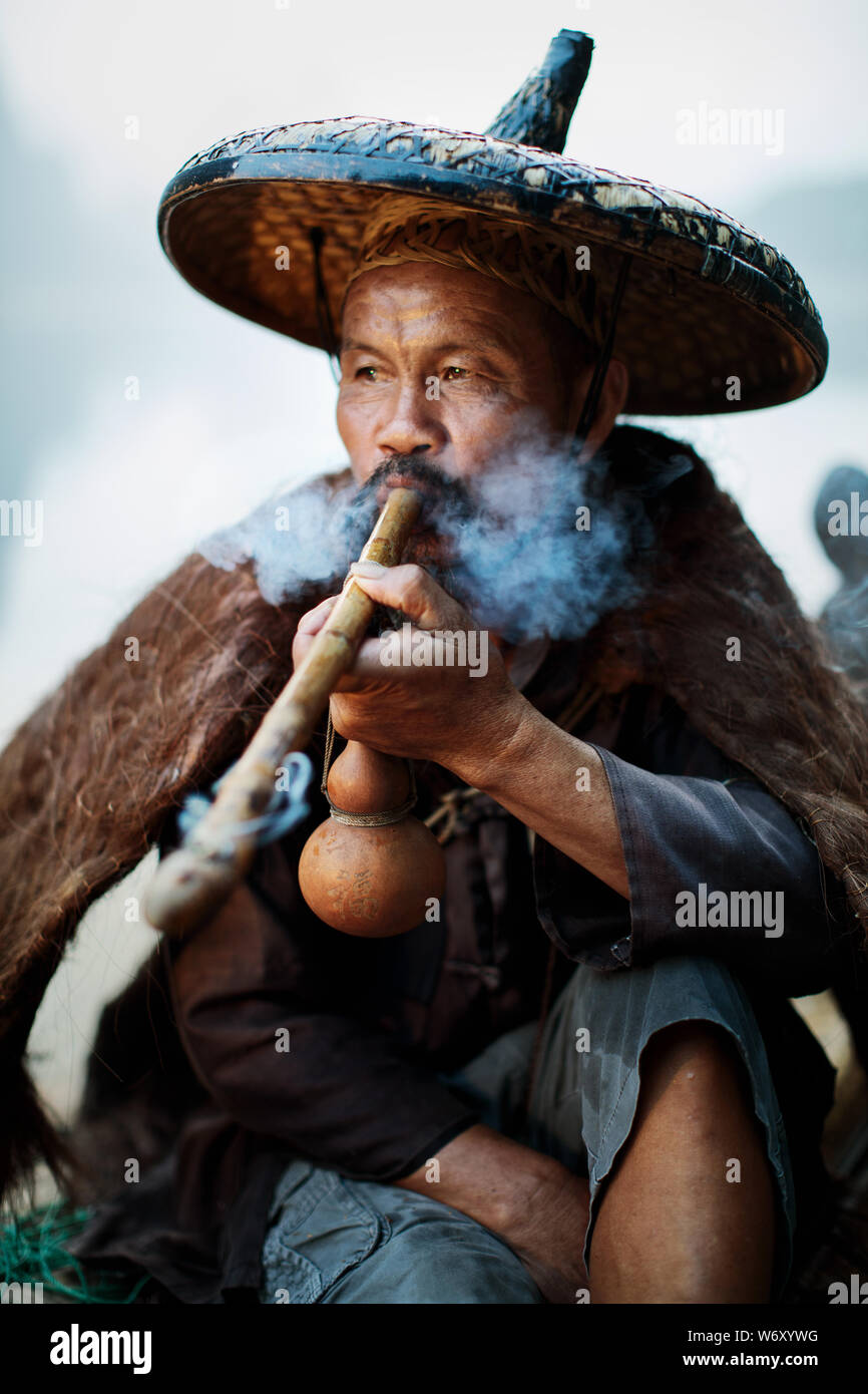 Cormorano tradizionale pescatore, Blackbeard, fumare la mattina presto in abiti tradizionali. Xingping, Cina. Foto Stock