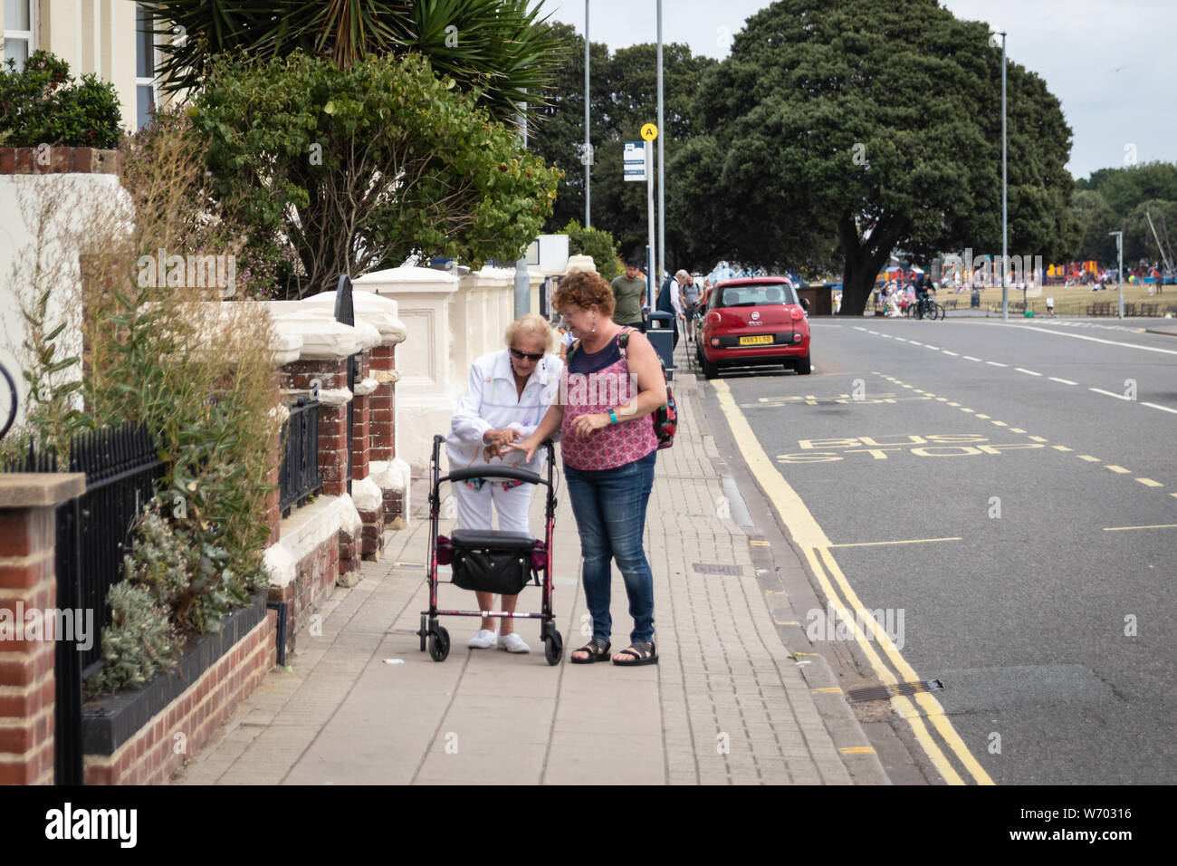 Una figlia cresciuta ad aiutare l'anziana madre a camminare usando un aiuto per la mobilità o Zimmer frame, i vecchi/persona disabile con accompagnatore Foto Stock