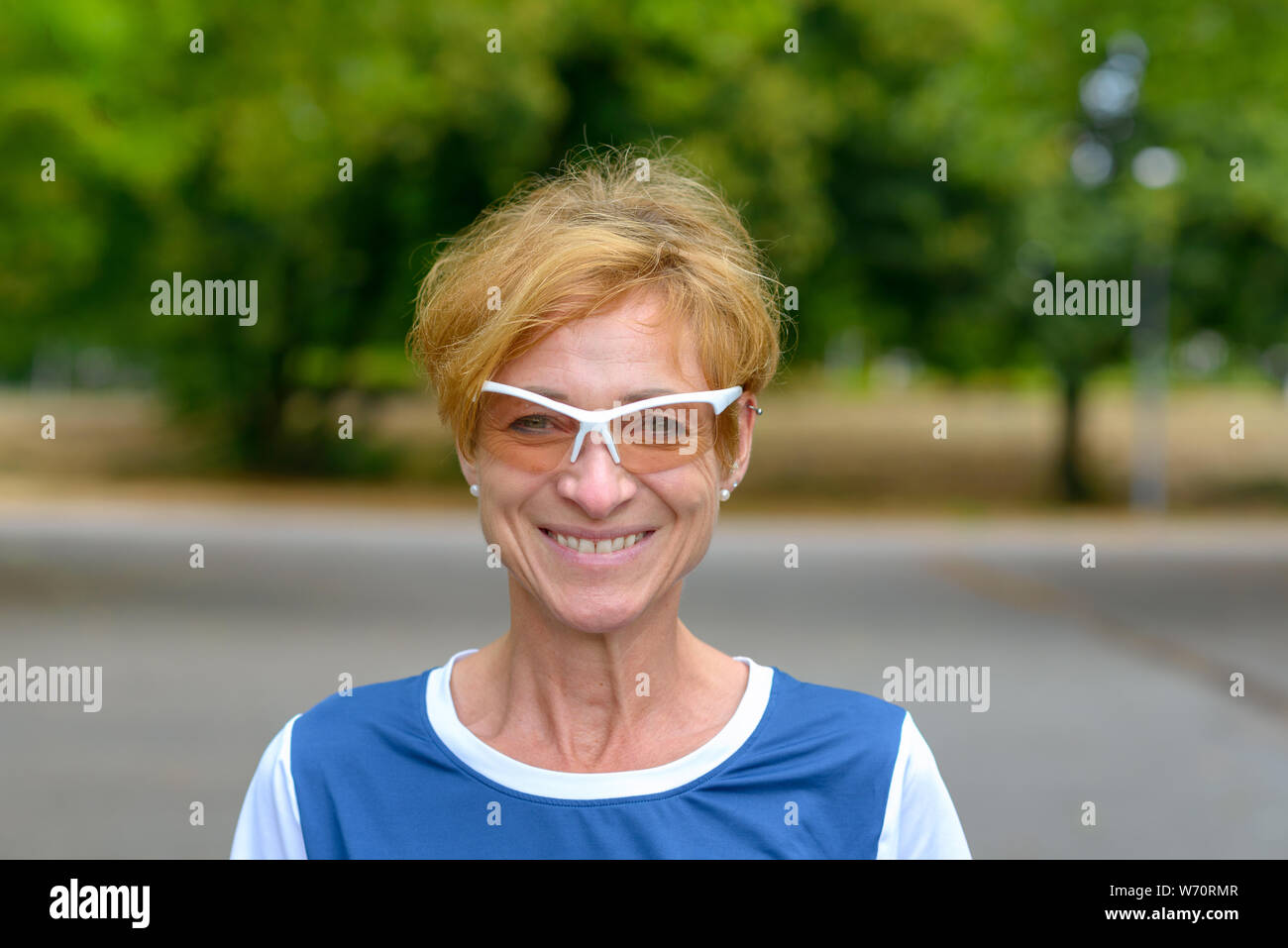 Felice donna vivace nel quartiere alla moda di occhiali moderni in piedi all'aperto su una tranquilla strada a guardare la fotocamera con un raggiante sorriso amichevole Foto Stock