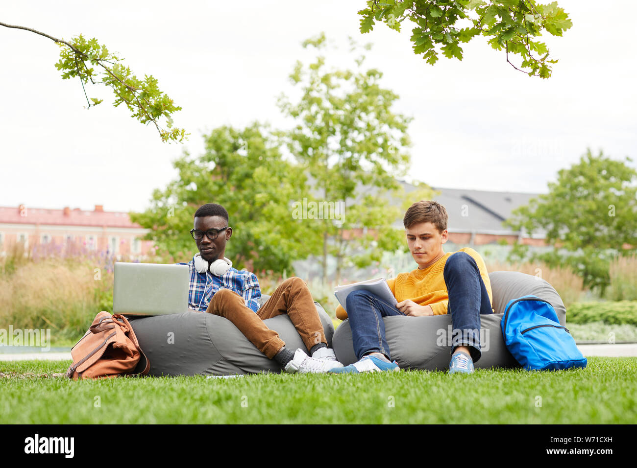 A piena lunghezza ritratto di due studenti internazionali che studiano all'aperto mentre è seduto sui sacchi di fagioli all'aperto in campus, spazio di copia Foto Stock