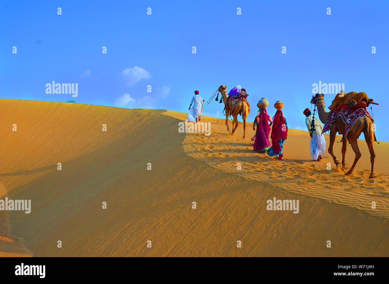 Due cameleers e le donne con i cammelli a piedi sulle dune di sabbia del deserto di Thar contro il cielo blu , Jaisalmer, Rajasthan, India Foto Stock