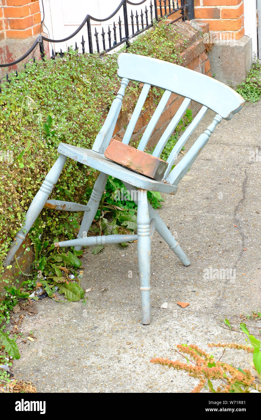 West Sussex, in Inghilterra, Regno Unito. La rottura di un dipinto di sedia in legno con una casa mattone sopra abbandonato in un giardino sul davanti. Foto Stock