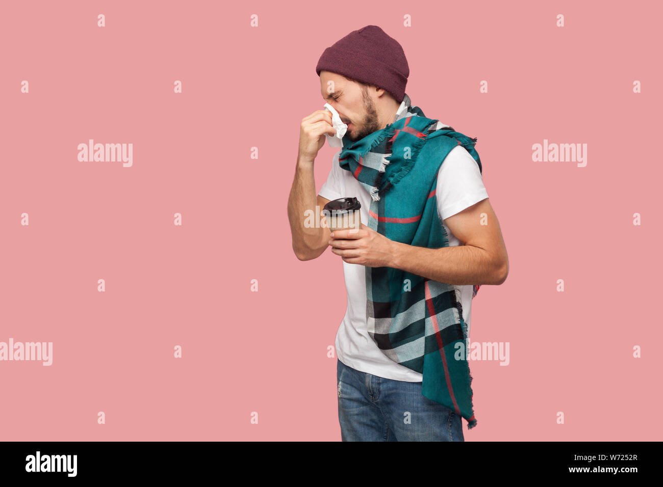 Ritratto di malattia moderna giovane barbuto hipster Uomo in camicia bianca e casual hat in piedi,starnutisce del tovagliolo, tenendo una bevanda calda nel bicchiere di carta. Piscina Foto Stock