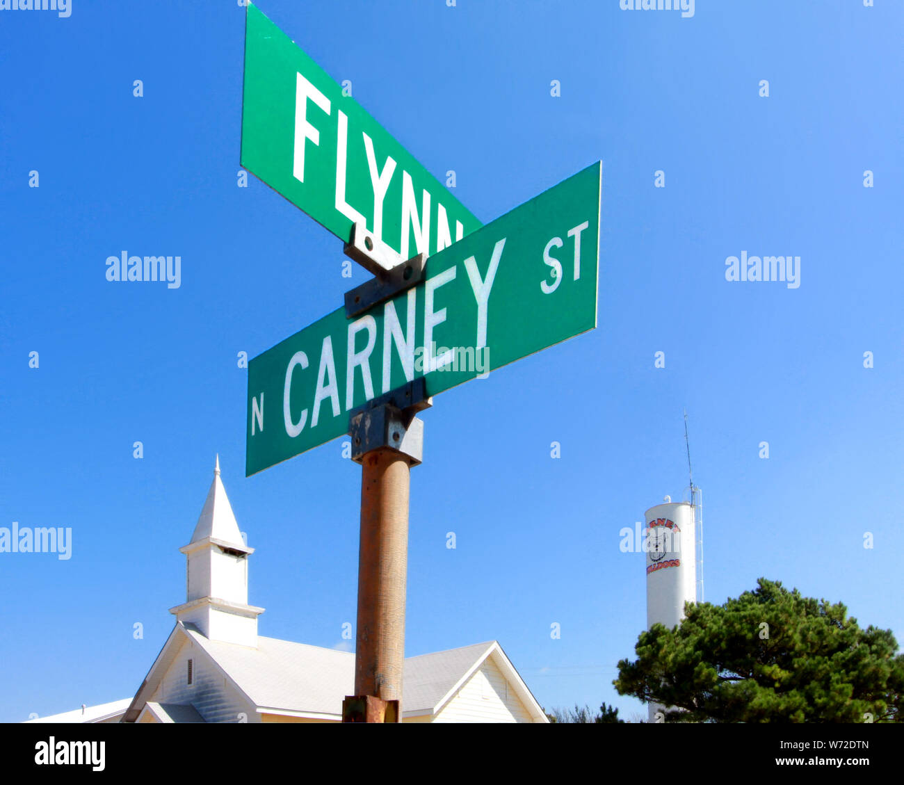 Una chiesa bianca il campanile e il Carney Bulldogs water tower sostare dietro il cartello stradale in corrispondenza della zona di spigolo di Flynn e Carney Street in Carney Oklahoma. Foto Stock
