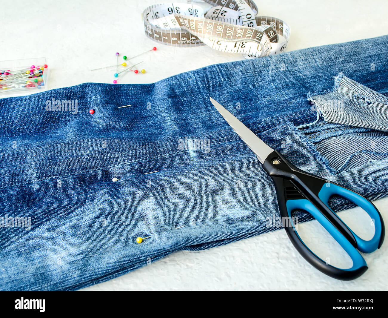 Jeans blu con foro grande piegato a metà, perni di cucitura, bianco di personalizzare il nastro con centimetri e pollici e forbici. Accorciare i jeans. Il DIY è in corto circuito Foto Stock