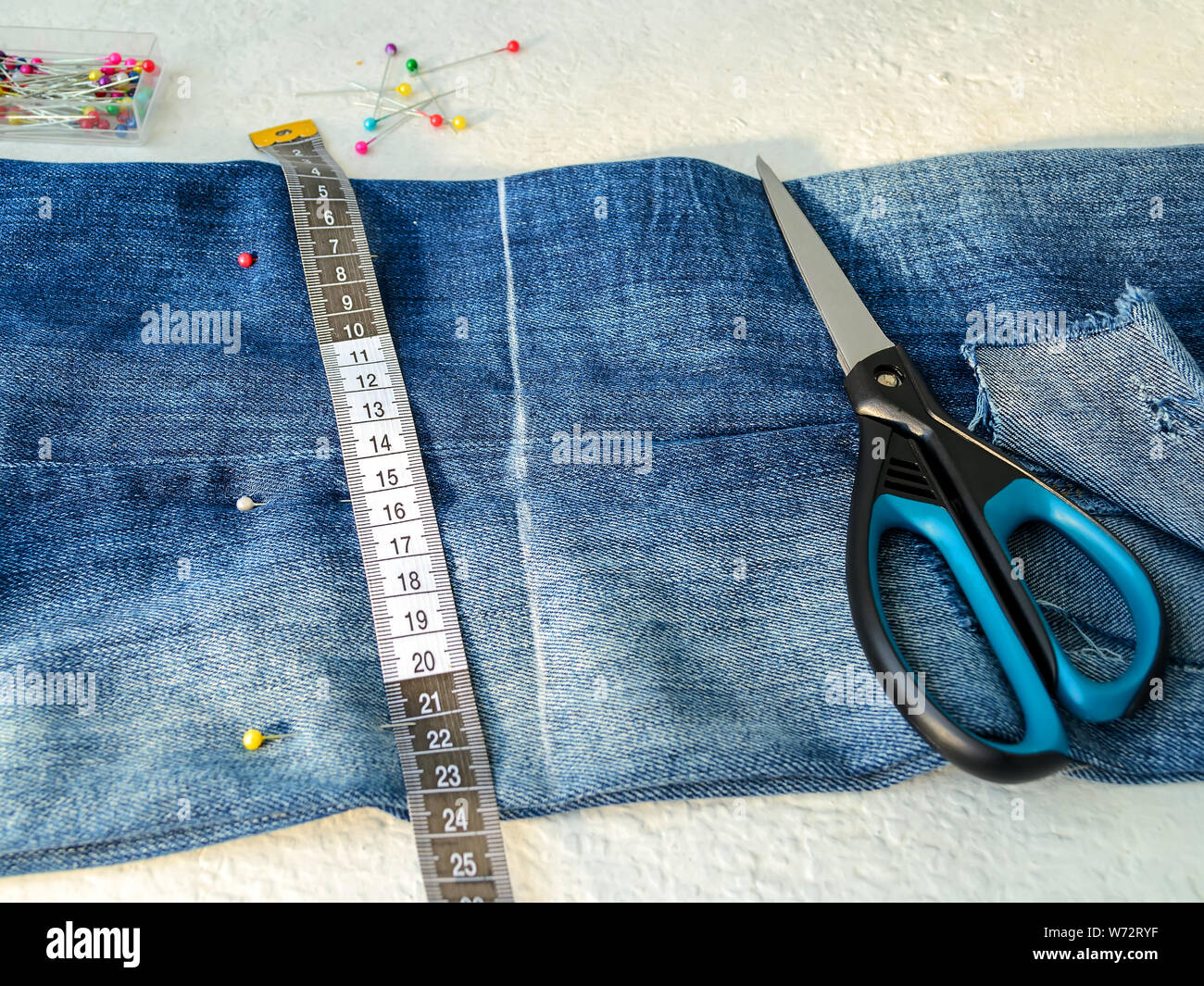 Jeans blu con foro grande piegato a metà, multi colore piedini di cucitura in una casella bianca del nastro su misura con centimetri e pollici e forbici. Accorciare th Foto Stock