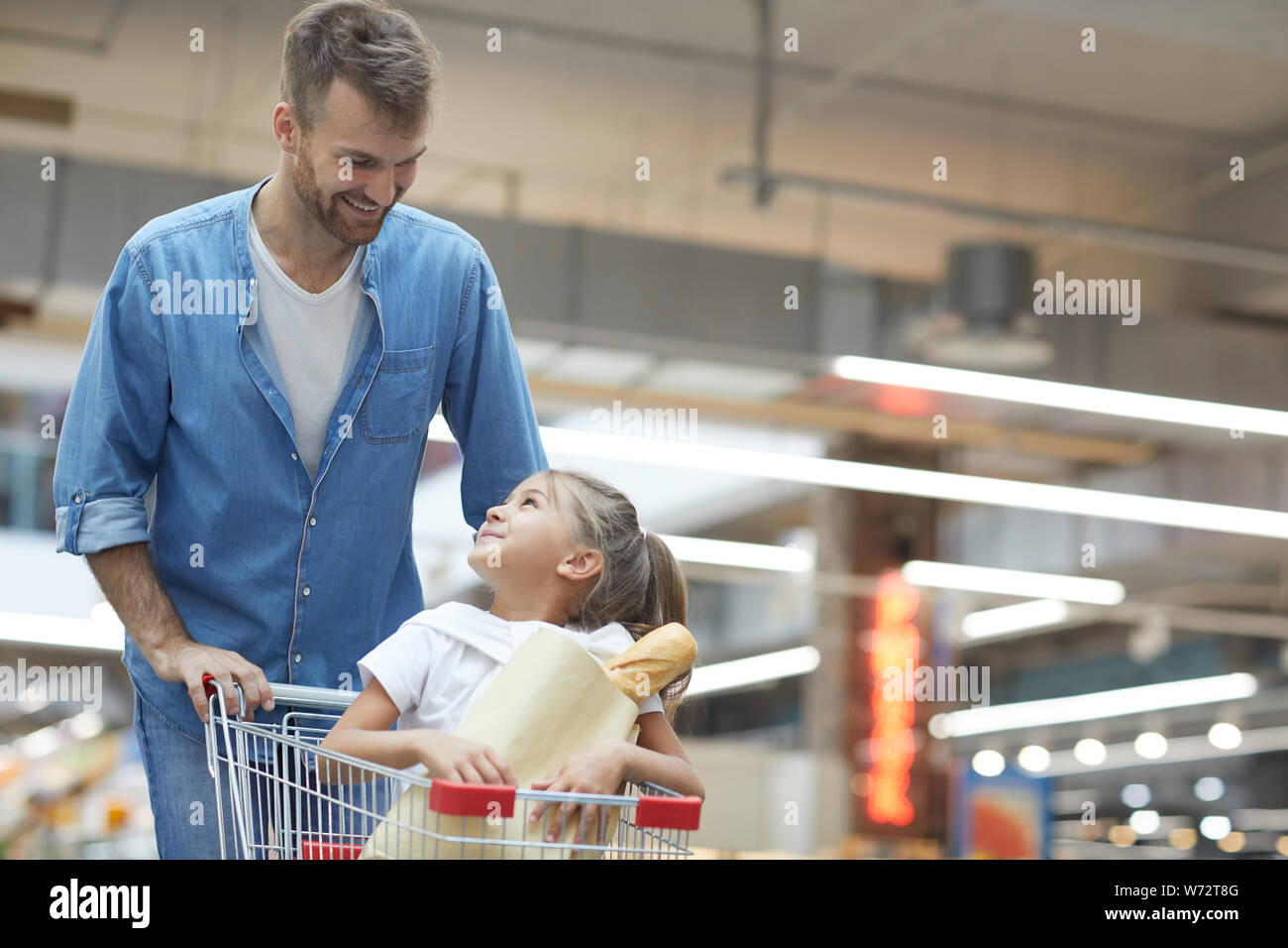 Ritratto di felice giovane padre a fare la spesa nel supermercato e sorridente a poco ragazza seduta nel carrello, spazio di copia Foto Stock
