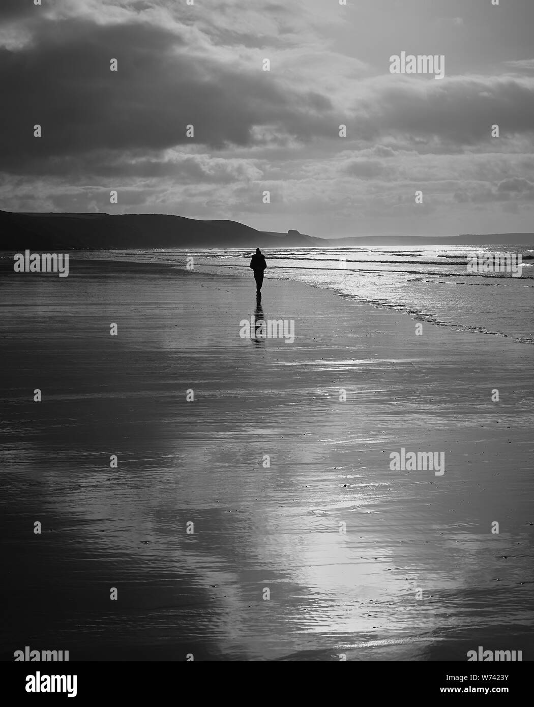 Solitudine:Una silhouette di un individuo a piedi lungo una spiaggia desolata in una mattinata d'inverno. L'immagine in bianco e nero mette in evidenza le riflessioni & scena. Foto Stock