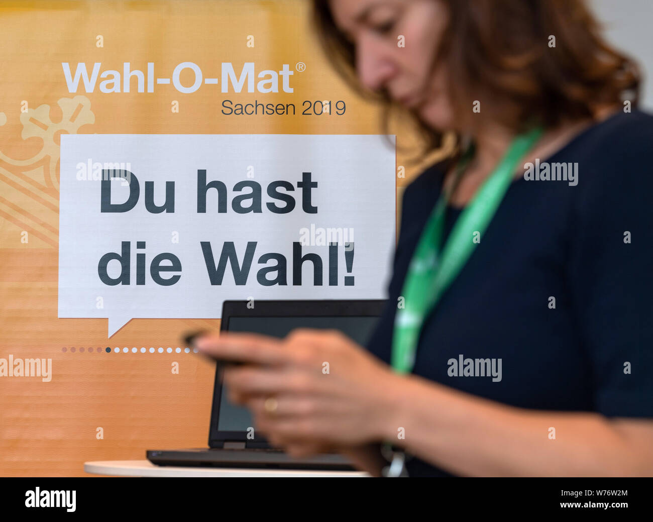 05 agosto 2019, Sassonia, Dresda: Pamela Brandt dall'Agenzia federale per l educazione civica sta con il suo smartphone davanti a un computer portatile e di un banner pubblicitario per la Wahl-O-Mat del Sassone Agenzia di Stato per l'educazione civica durante la presentazione in stato sassone in Parlamento. Con il Wahl-O-Mat, gli utenti possono informarsi circa le posizioni delle parti in esecuzione per il 2019 alle elezioni statali sulla base di 38 tesi di laurea su vari settori politici. Foto: Robert Michael/dpa-Zentralbild/dpa Foto Stock