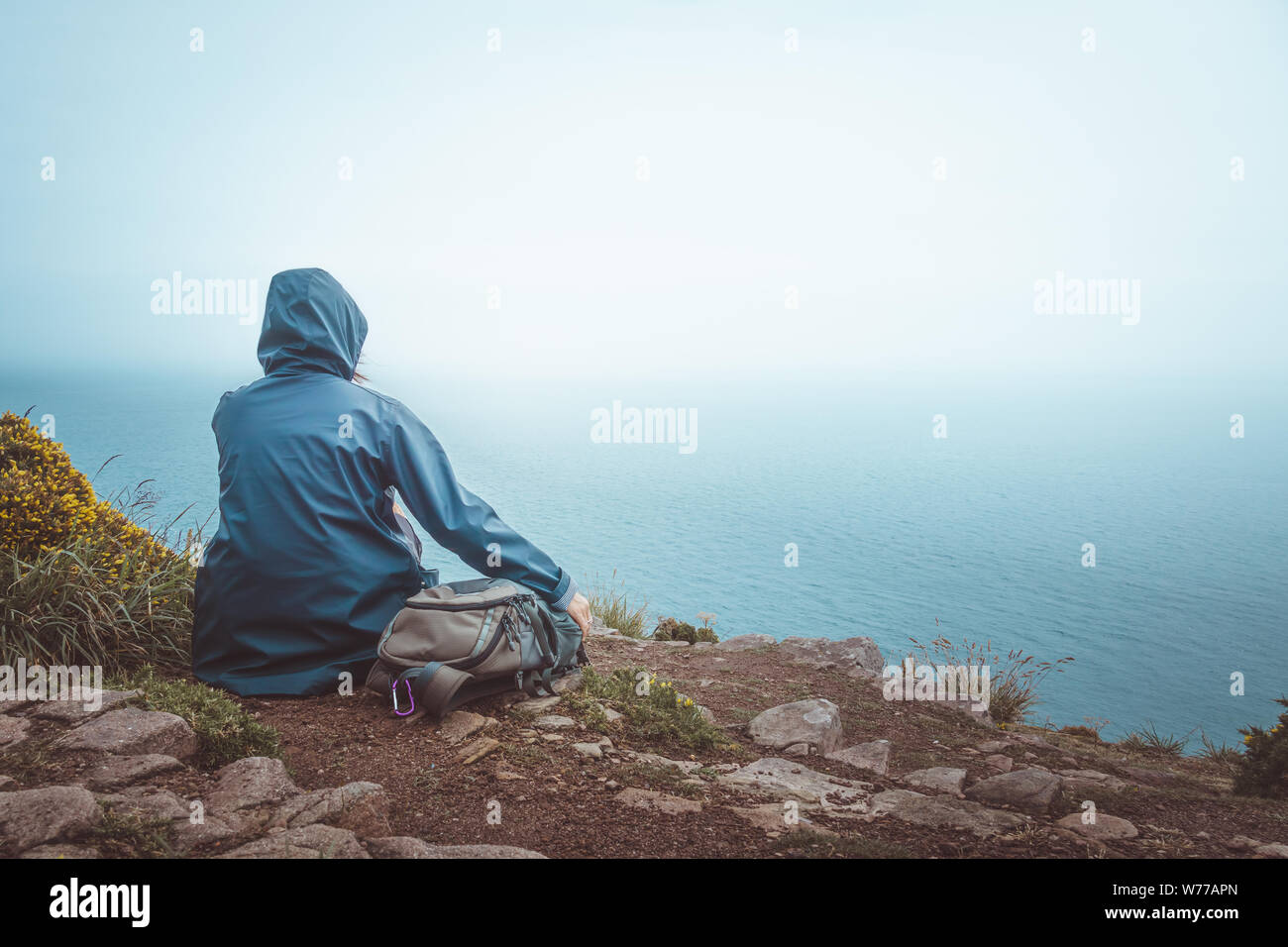 Vista posteriore di un escursionista donna seduta sul bordo di una scogliera con un impermeabile blu e zainetto guardando l'orizzonte e il mare. Foto Stock