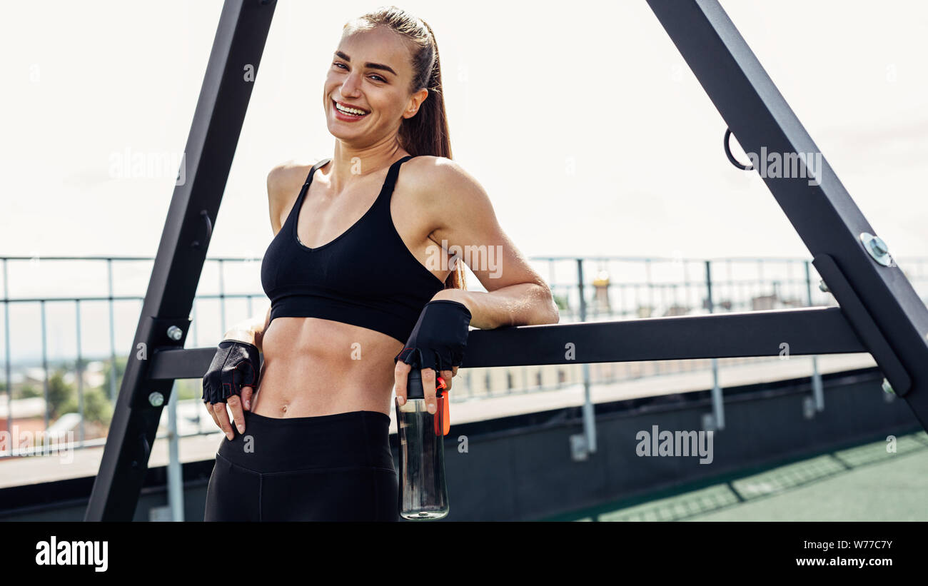 Donna sorridente in sportswear appoggiata sul power rack tenendo una bottiglia con acqua Foto Stock