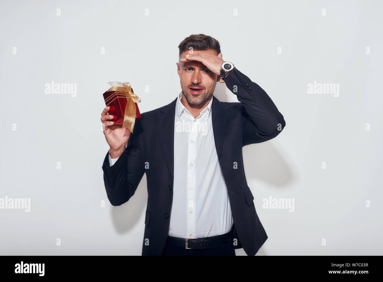 Ho qualcosa per voi buon uomo d'affari bearded osservante in vestito classico che tiene una scatola del regalo e che guarda la macchina fotografica mentre si levano in piedi contro sfondo grigio. Presente. Vacanze Foto Stock
