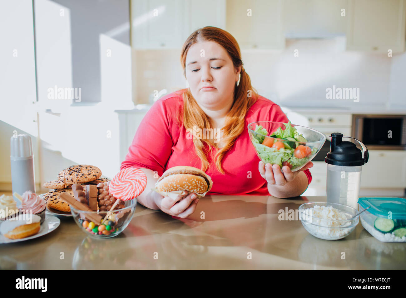 Fat giovane donna in cucina e salotto a mangiare cibo. Uno stile di vita sano. Guardando il burger e ciotola insalata. Corpo positivo. Caramelle sulla sinistra. Pasto sano o Foto Stock