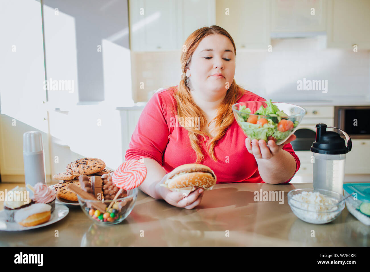 Fat giovane donna in cucina e salotto a mangiare cibo. Ciotola con insalata in mano. Plus size model look a. Corpo positivo. Confuso giovane donna in kitche Foto Stock